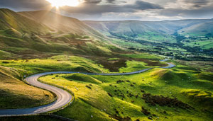 A winding road through lush green hills under a dramatic sky, with sunlight filtering through the clouds, showcasing a picturesque landscape.