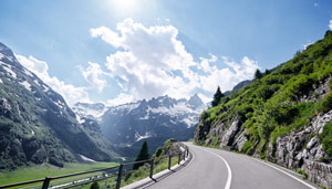 Curving road through lush green mountains under a bright blue sky with fluffy clouds and snow-capped peaks.