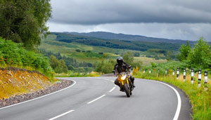 A motorcycle rider navigating a curved road through a scenic landscape with green hills and overcast skies.