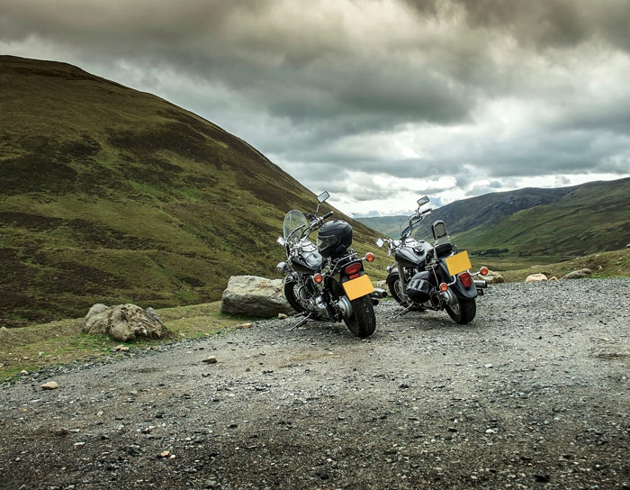 Two motorcycles parked on a gravel road with mountains in the background, showcasing a scenic landscape perfect for motorcycle adventures.