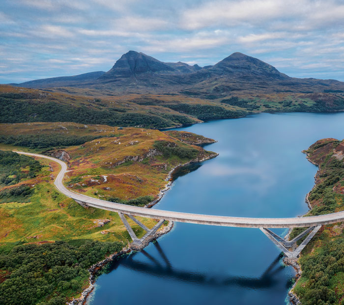 Aerial view of a bridge spanning a lake surrounded by green hills and mountains under a cloudy sky, showcasing scenic landscape beauty.