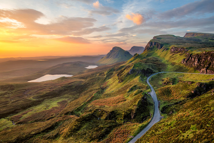 A scenic landscape featuring rolling green hills, a winding road, and a serene lake under a colorful sunset sky. The majestic mountains in the background create a breathtaking backdrop for this picturesque view of nature.