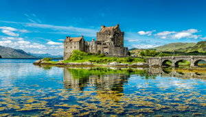 A picturesque view of a medieval castle surrounded by a serene lake and lush greenery under a bright blue sky, reflecting the structure and landscape in the water, ideal for landscape photography enthusiasts.