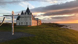 A scenic view of a historic building with a unique architectural style, surrounded by lush green grass and overlooking the ocean at sunset, with a colorful signpost nearby. Explore coastal beauty.