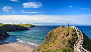 A scenic coastal view featuring a person standing on a wooden walkway overlooking a sandy beach and clear blue water with rolling green hills in the background, perfect for visual inspiration and relaxation.