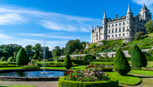 A picturesque landscape featuring a grand castle surrounded by manicured gardens, water fountains, and vibrant flowers under a clear blue sky.