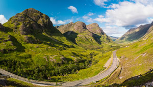 A panoramic view of a lush green valley surrounded by majestic mountains under a bright blue sky, with a winding road cutting through the landscape, showcasing the natural beauty of the area. Scenic travel.