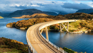 Curved bridge over a tranquil body of water with mountains in the background, showcasing scenic beauty.