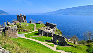 Scenic view of an ancient castle ruins along a lush green hillside, overlooking a tranquil lake and distant mountains under a clear blue sky. Ideal for exploring Scottish history.